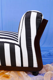 Close up of retro Amanta 24 chair with black and white striped upholstery and a brown frame against a blue backdrop with orange chiffon floor covering. 