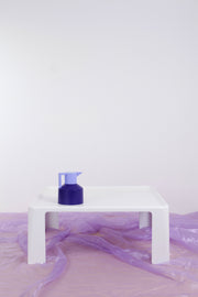 White Amanta coffee table against white backdrop and purple chiffon floor covering. There is a two-tone purple coffee pot on top of the retro coffee table. 