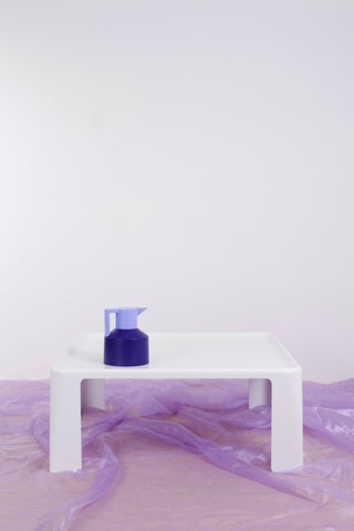 White Amanta coffee table against white backdrop and purple chiffon floor covering. There is a two-tone purple coffee pot on top of the retro coffee table. 