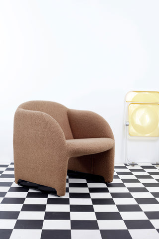 Vintage caramel Pierre Paulin 'Ben' chair against a white background and a chequered floor. A Folded yellow Plia chair rests against the wall. 