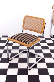 Retro Bauhaus Breuer Cesca cantilevered chair with caned back and upholstered seat against a white backdrop and chequered flooring. A folded Plia chair can be seen in the background.  