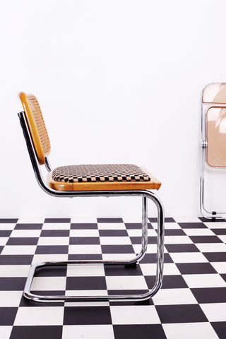 Vintage Breuer Cesca chair with caned back and cheque upholstered seat against a white background and a chequered floor. A folded pink Plia chair leans against then rear wall.  