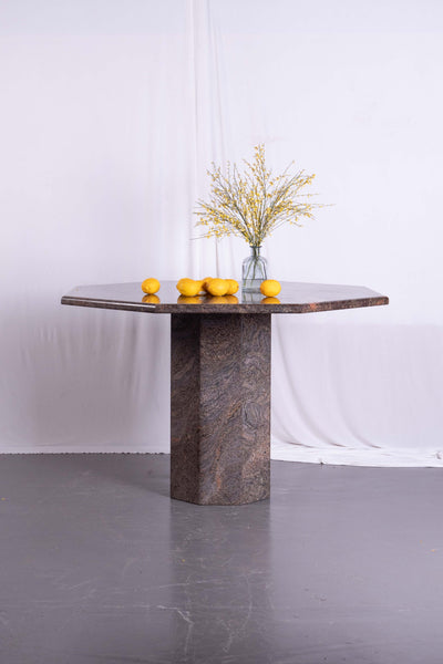 Octagonal marble dining table. On top of the table is a vase with yellow flowers. White draped fabric in the background