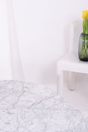 Leaf-shaped Carrara marble coffee table against white drapes with white plastic chair and vase filled with daffodills in the background.