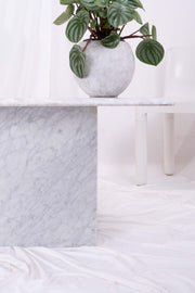 Close up shot of the base of this vintage Italian white marble coffee table. A plant sits on the tabletop; a white plastic chair is visible behind. The image is set in front of white drapes.