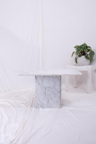 Vintage square Carrara marble coffee table with plant in concrete pot on top. In the background is a white bartoli chair and it is all set in front of white drapes.