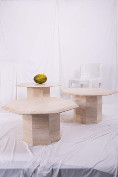 Set of three travertine nesting tables. On one of the coffee tables there is a melon. There is white fabric drapped and a plastic kartell chair in the background
